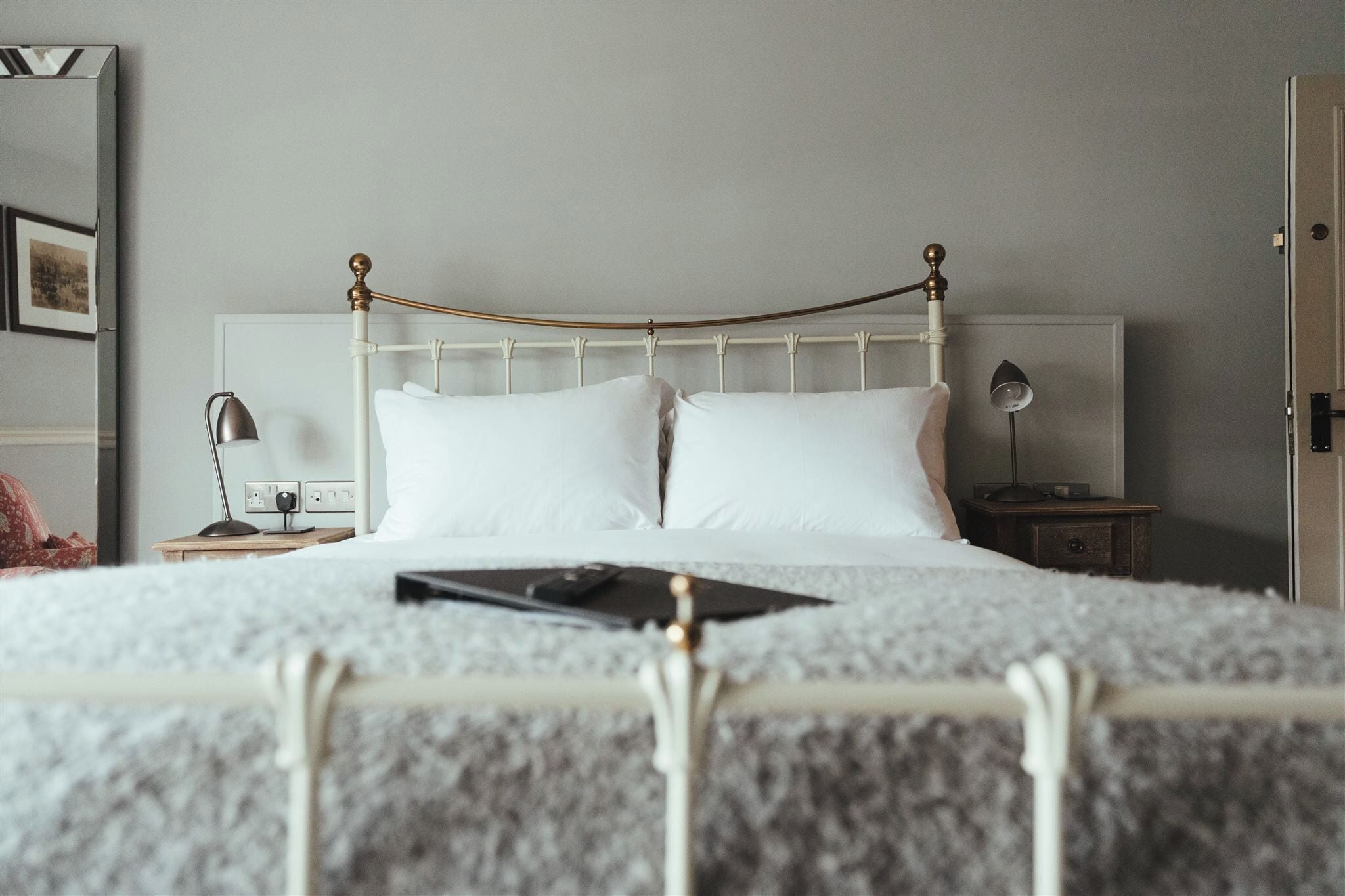 Una cama bien hecha con ropa blanca de cama y cabecera de metal, una mesita de noche con una lámpara y un cuadro en la pared.
