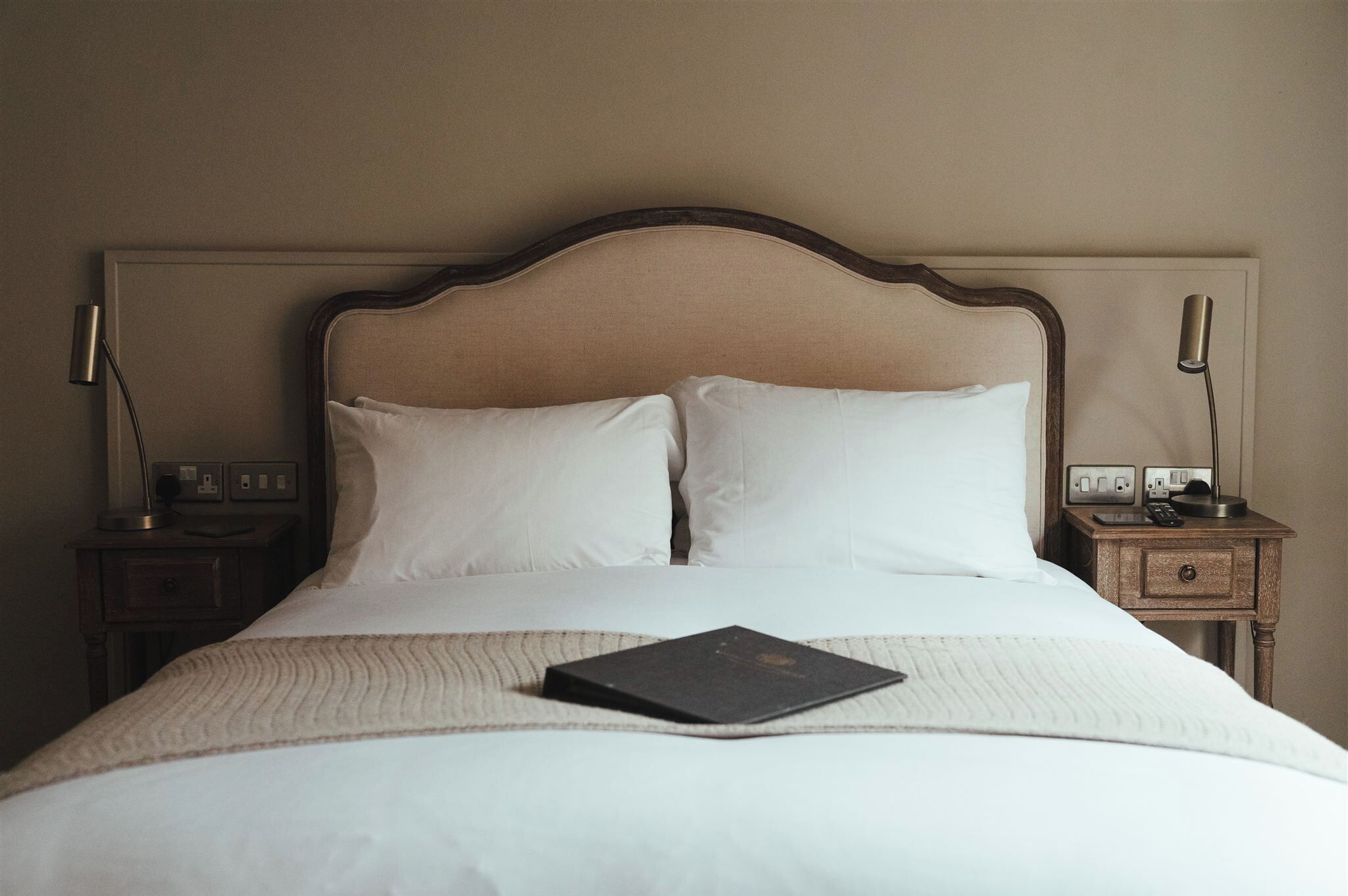 Una cama bien hecha con ropa de cama blanca, un libro oscuro sobre la cama y dos mesitas de noche con lámparas y una radio.