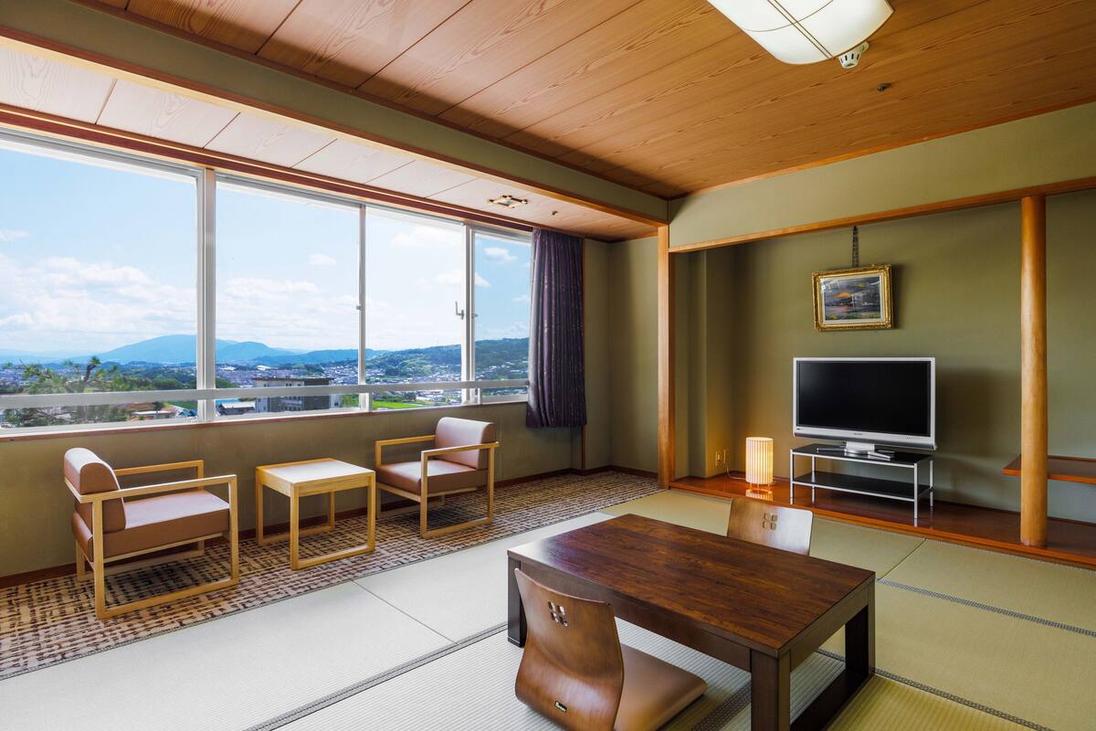 A hotel room with tatami flooring, a wooden ceiling, a large window with a view of mountains, a television, and a traditional Japanese-style sofa and table.