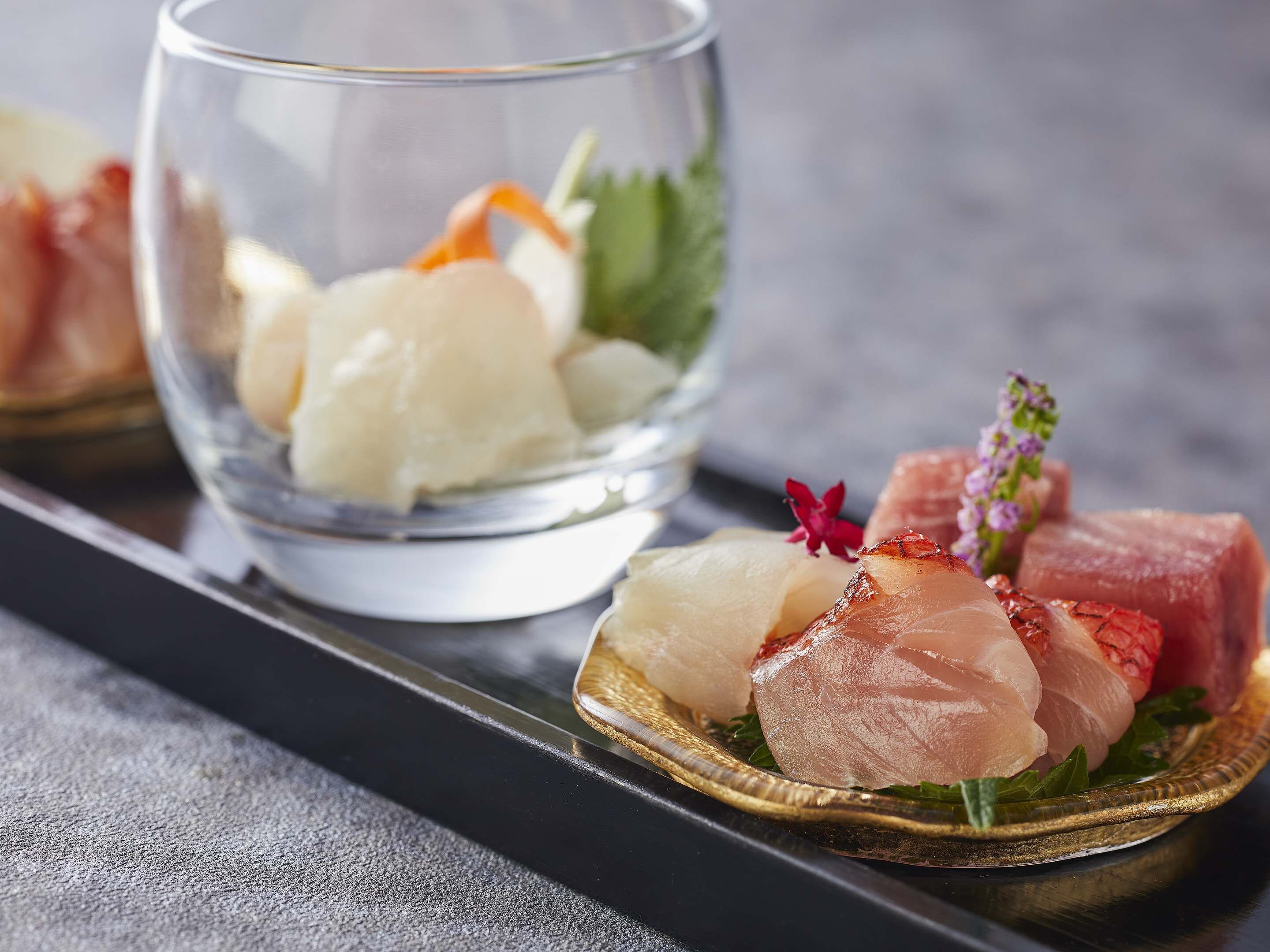 A glass with a seafood dish, a plate with sashimi, and a decorative flower.
