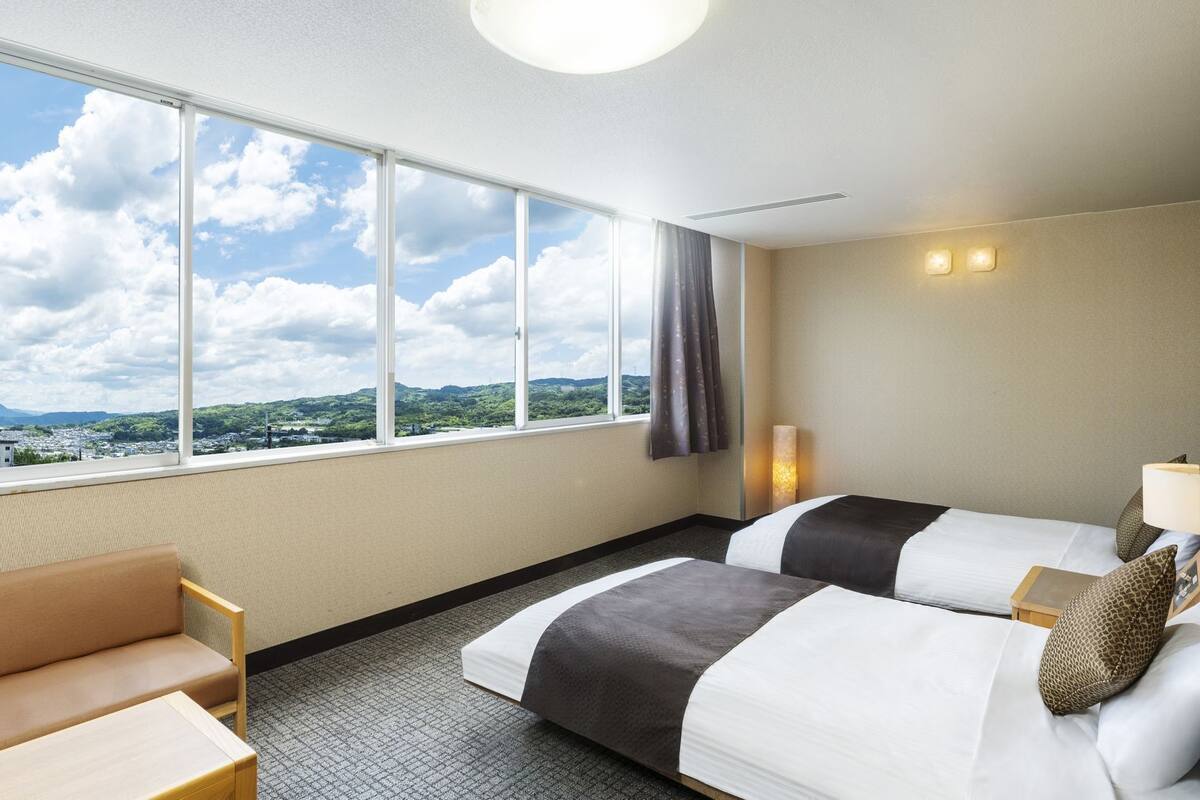 Ein Hotelzimmer mit zwei Betten, einem Schlafsofa, einem großen Fenster mit Blick auf Stadt und Berge sowie einer Deckenleuchte.