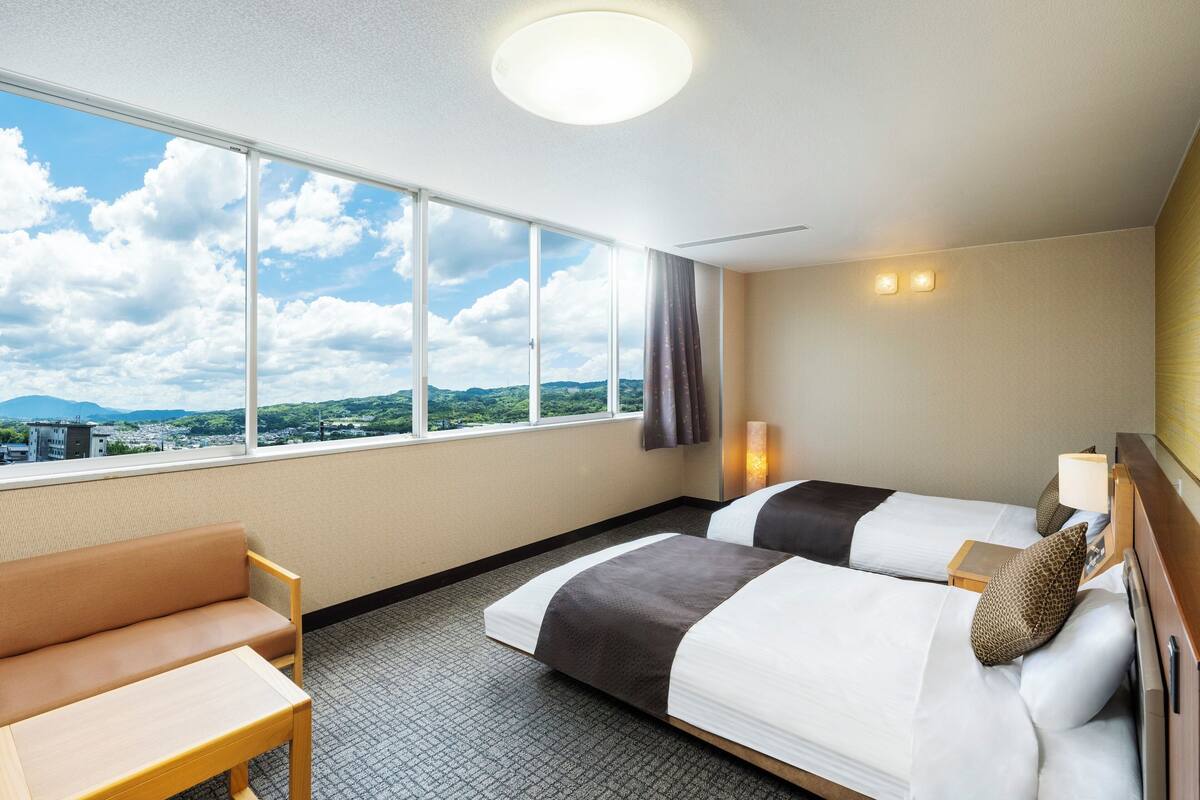 A hotel room with two beds, a sofa, a large window with a city view, and a ceiling light.