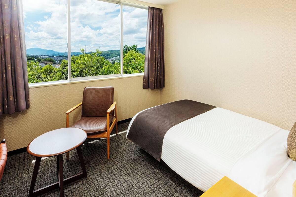 A hotel room with a bed, a chair, a small table, and a window offering a view of greenery and mountains.