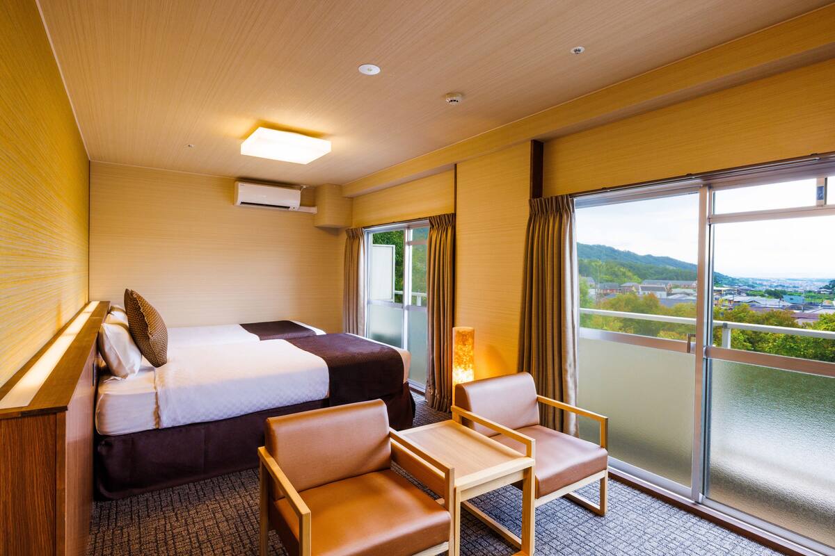 A hotel room with a bed, two chairs, a small table, and a large window offering a view of the landscape.