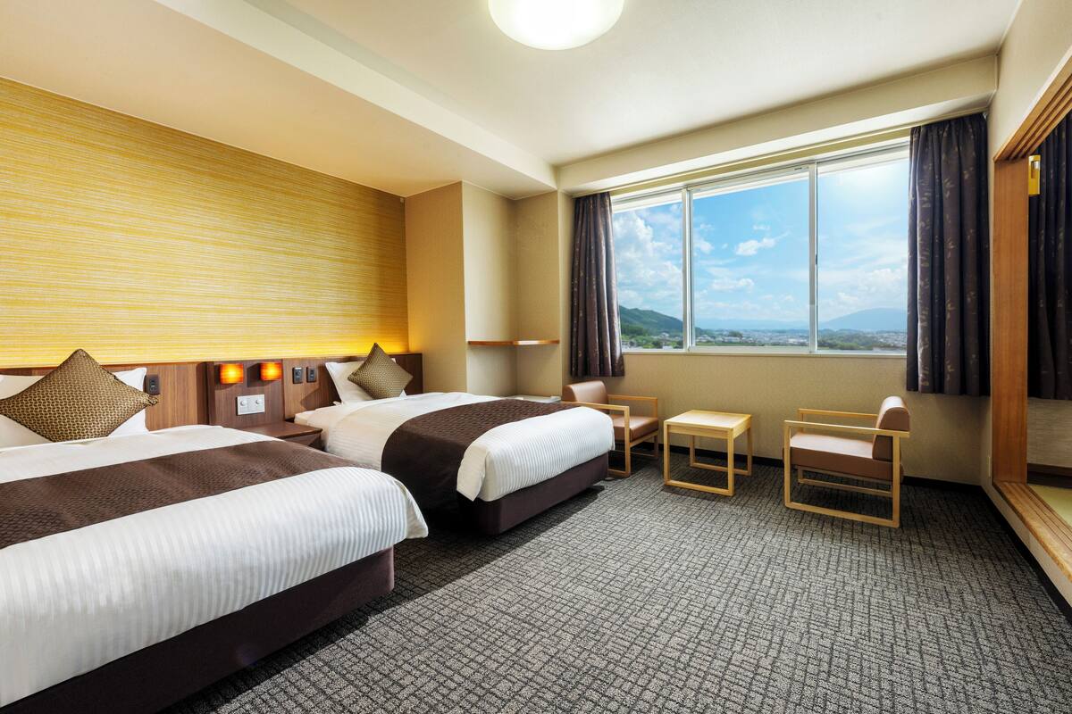 A hotel room with two beds, a small table, a chair, and a large window with a view of the mountains.