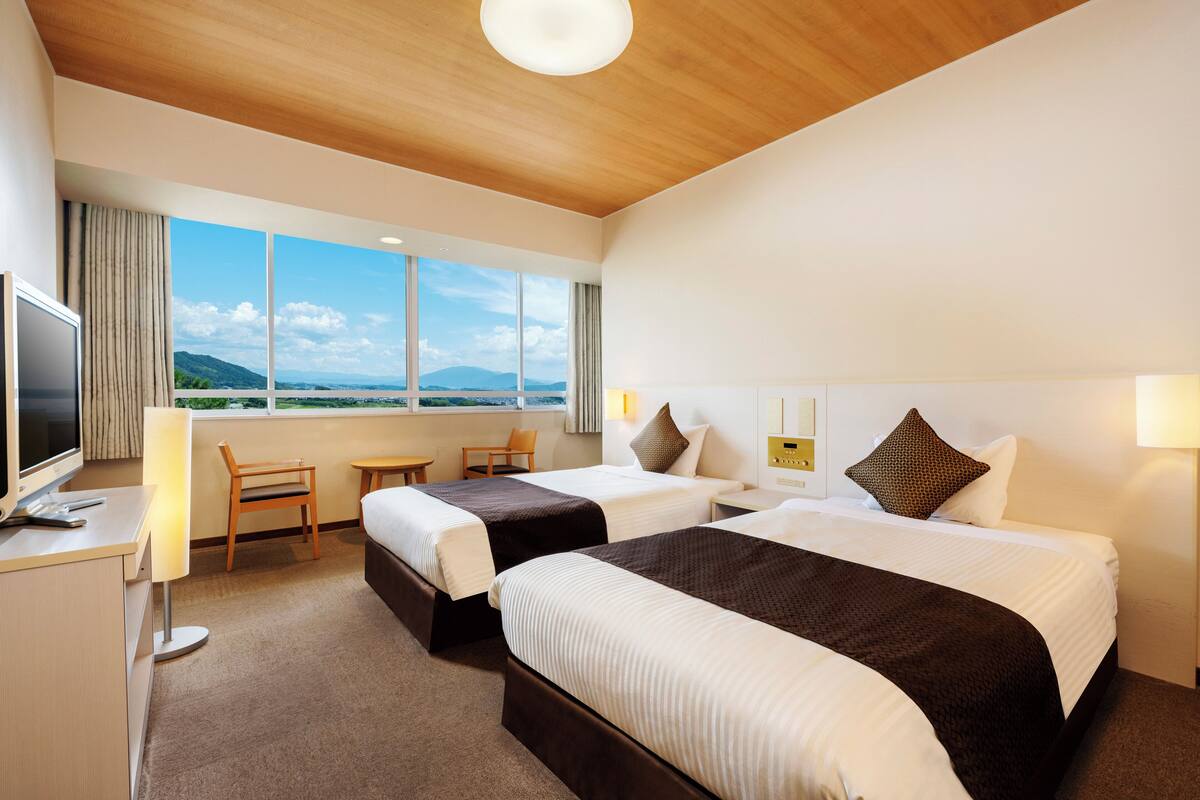 A hotel room with two beds, a desk with a TV, a chair, and a view of the mountains.