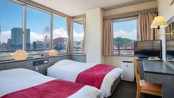 A hotel room with two beds, a desk, a TV, and a view of the cityscape.