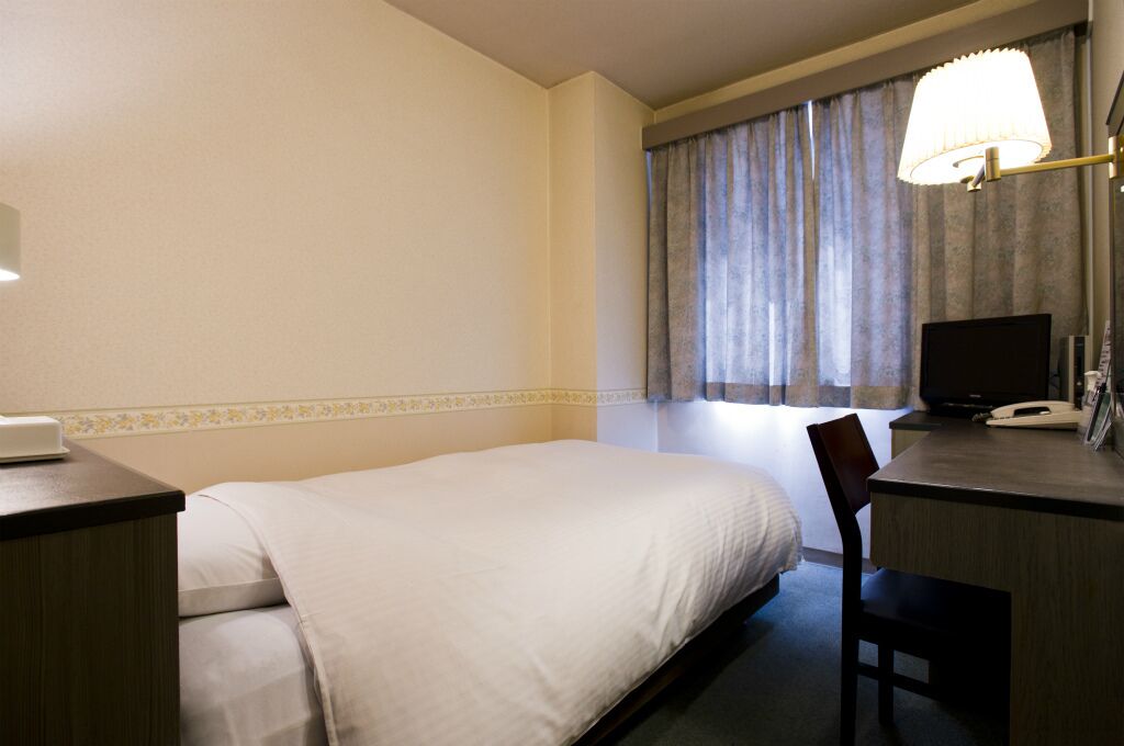 A hotel room with a bed, desk, chair, lamp, TV, and curtains.