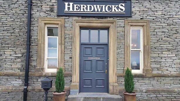 A stone building with a sign reading "Herdwicks" above a dark blue door labeled "HODDBARROW". - Herdwicks (Millom)