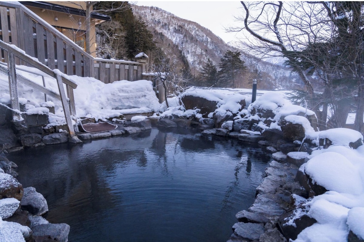 A traditional Japanese onsen with a wooden staircase leading to a stone bath surrounded by snow-covered rocks and trees.