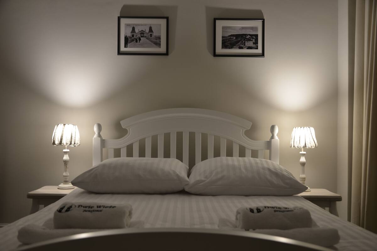 Een net opgemaakt bed met witte lakens en twee bedlampjes. Boven het bed hangen twee ingelijste foto's.