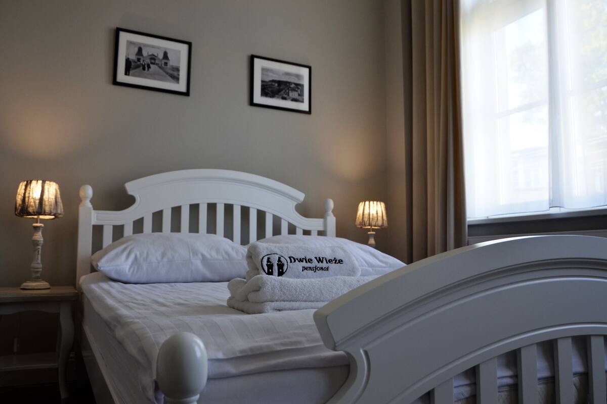 Een net opgemaakt bed met een handdoek waarop staat 'Dwie Wieże pensjonat', twee ingelijste foto's aan de muur, en een raam met gordijnen.