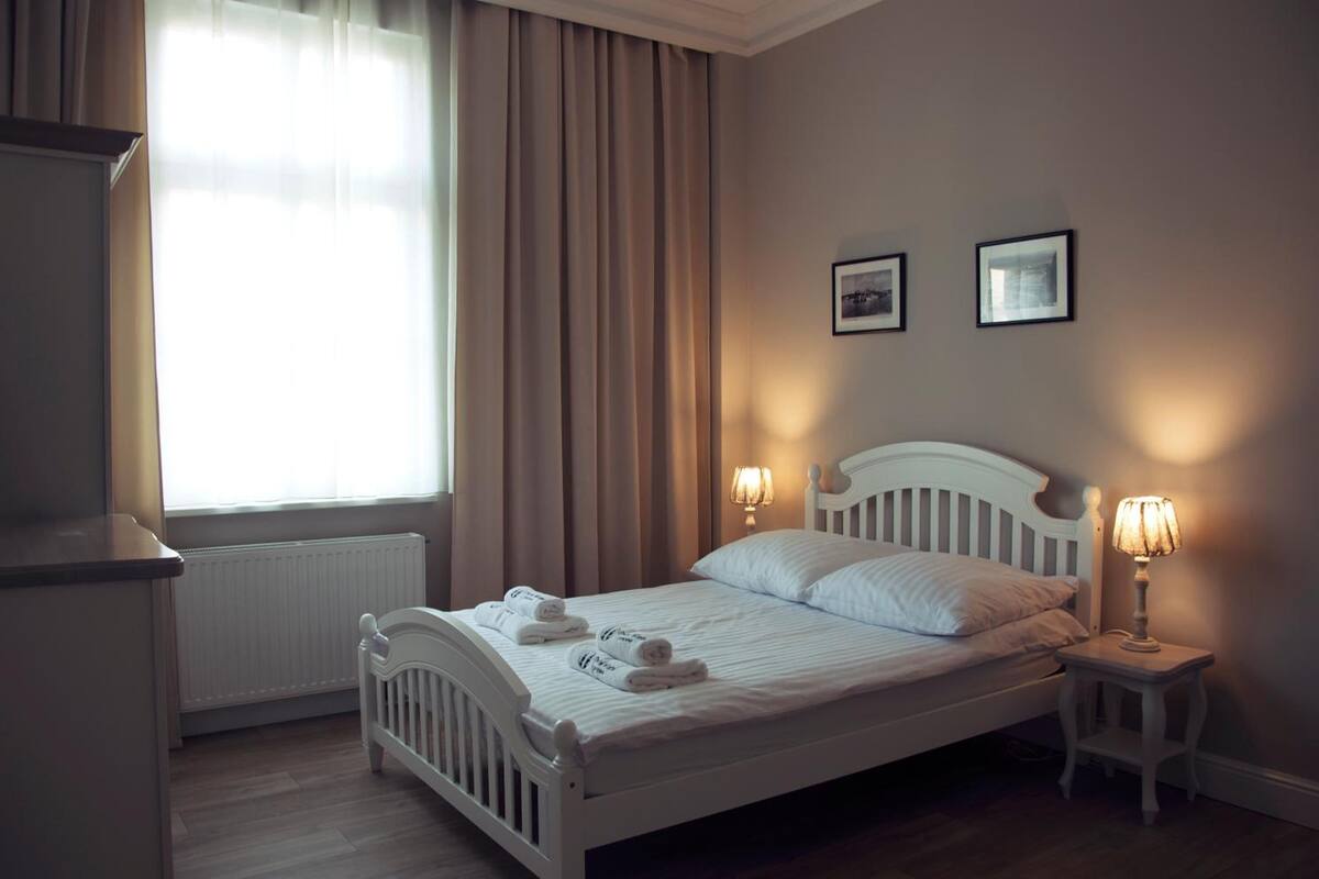 Een net opgemaakt bed met twee bedlampen, een radiator en een raam met gordijnen.