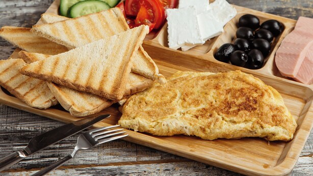 Ein Holztisch mit einem Frühstücksbrett, das Toast, Käse, Oliven, Tomaten, Gurken und Schinken beinhaltet. Außerdem sind ein Teekessel und Tassen zu sehen.