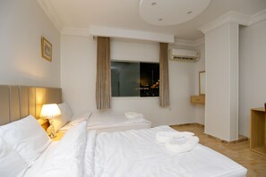 A hotel room with two beds, a nightstand with a lamp, a window with curtains, an air conditioning unit, and a wooden cabinet. - Makadi Pyramids View (Giza)