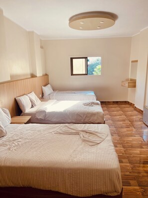 A hotel room with two beds, a wooden floor, a window with a view, and a ceiling light fixture. - Makadi Pyramids View (Giza)
