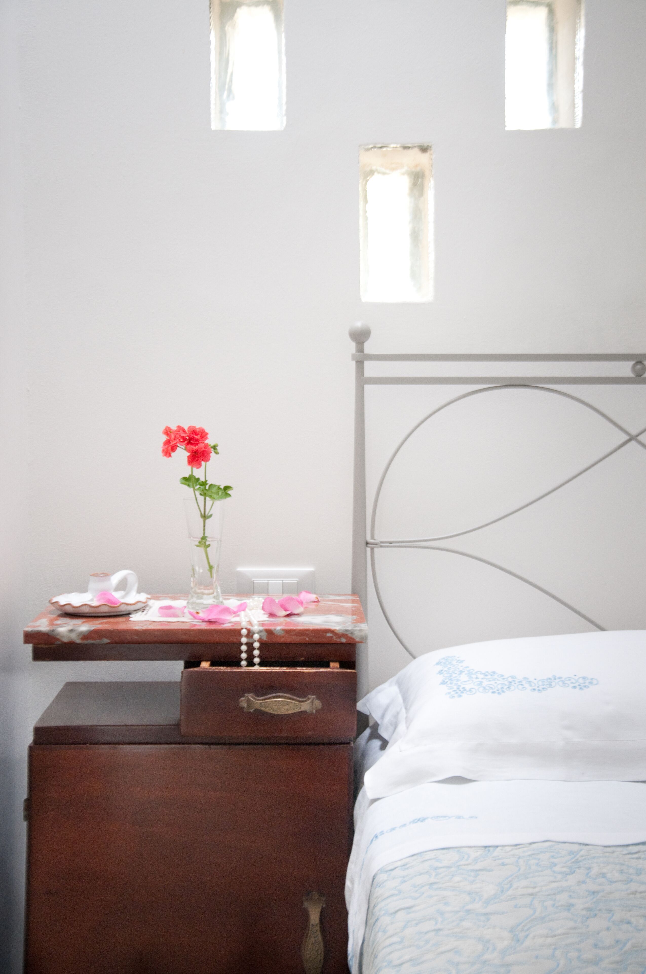 Une chambre à coucher avec un lit, une commode, une table de chevet, un vase avec des fleurs et une fenêtre.