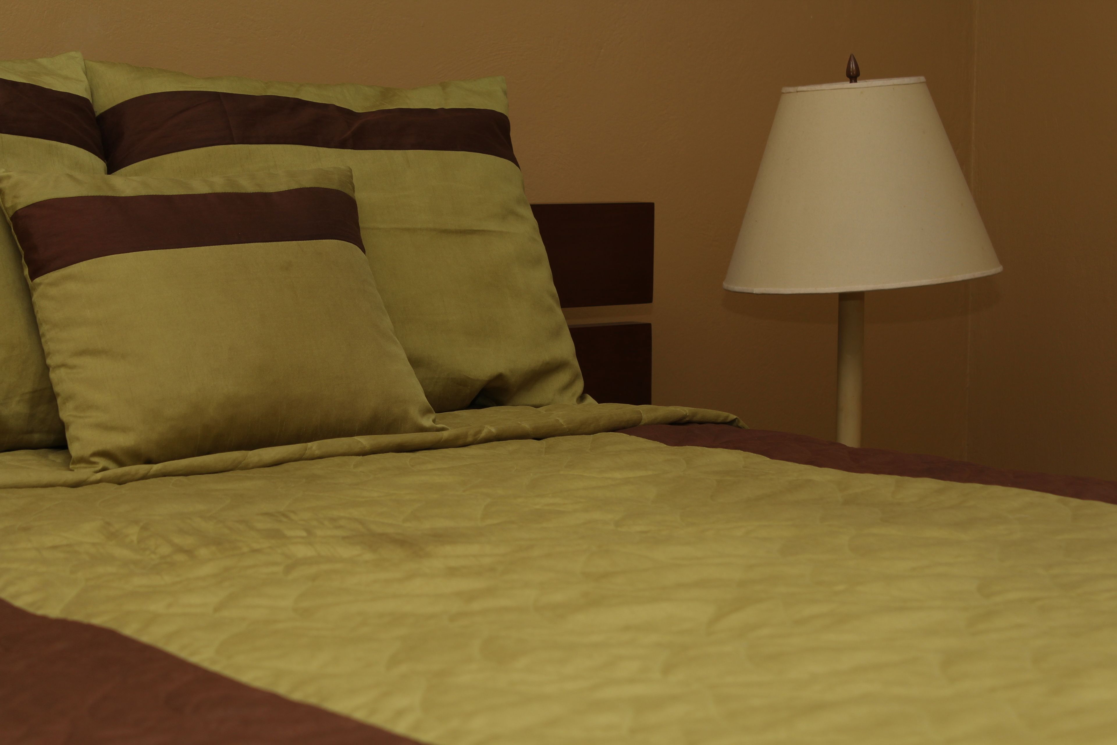 Un lit avec une literie aux couleurs vert et marron, une lampe de chevet blanche et un mur marron.
