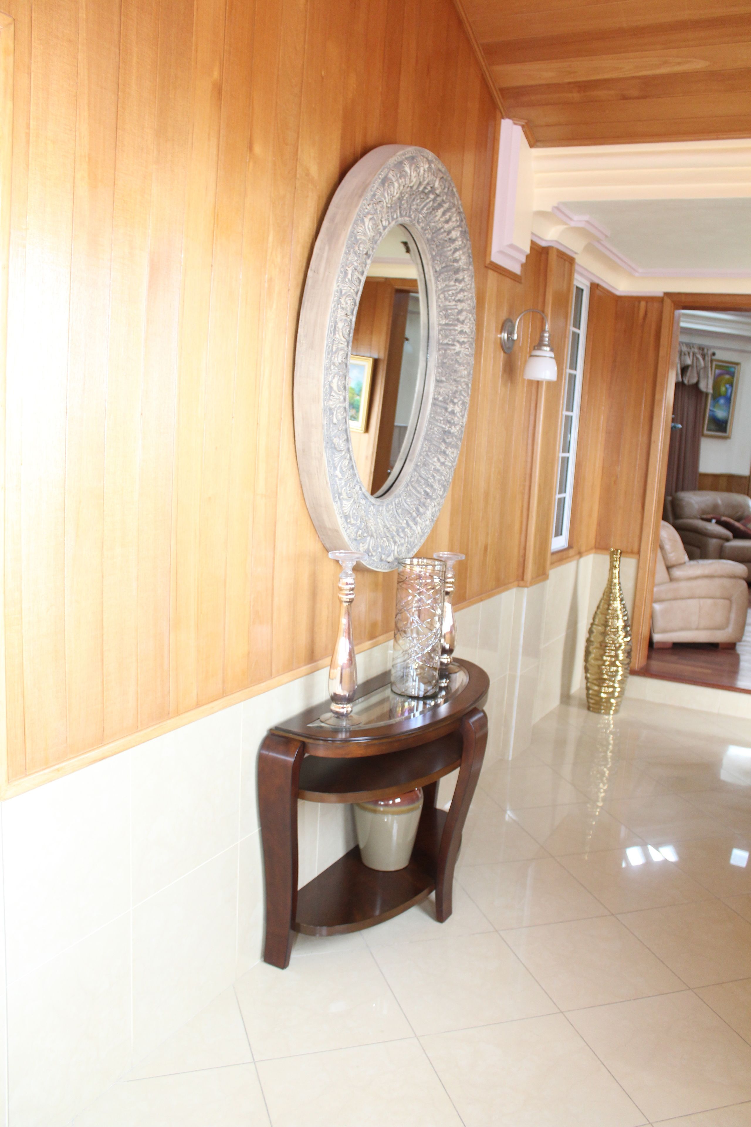 A wooden-paneled hallway with a round mirror on a console table, a vase, and a lamp.