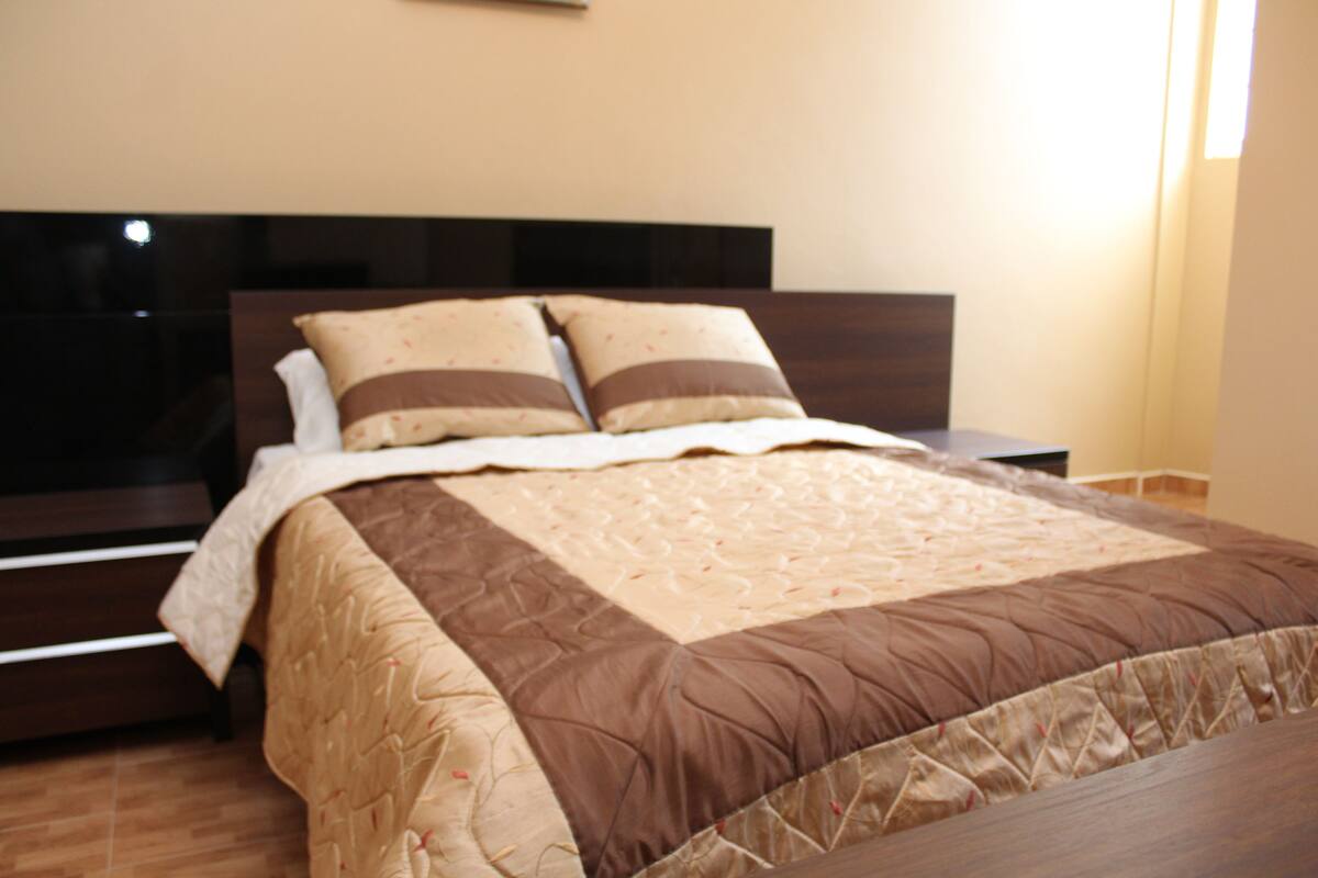 A neatly made bed with a brown and beige quilt, a dark headboard, and a wooden nightstand beside it.
