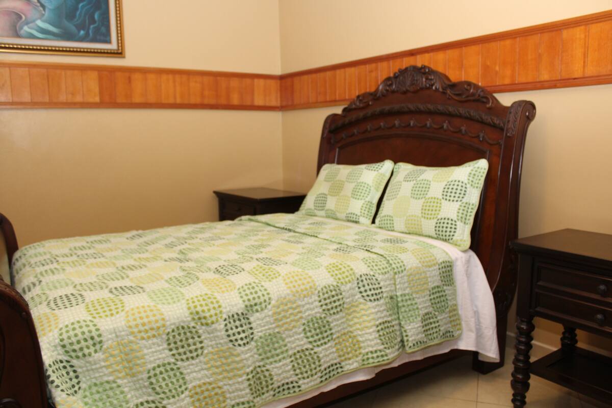 A bedroom with a wooden bed, a green and white patterned bedspread, a nightstand, and a wooden headboard.