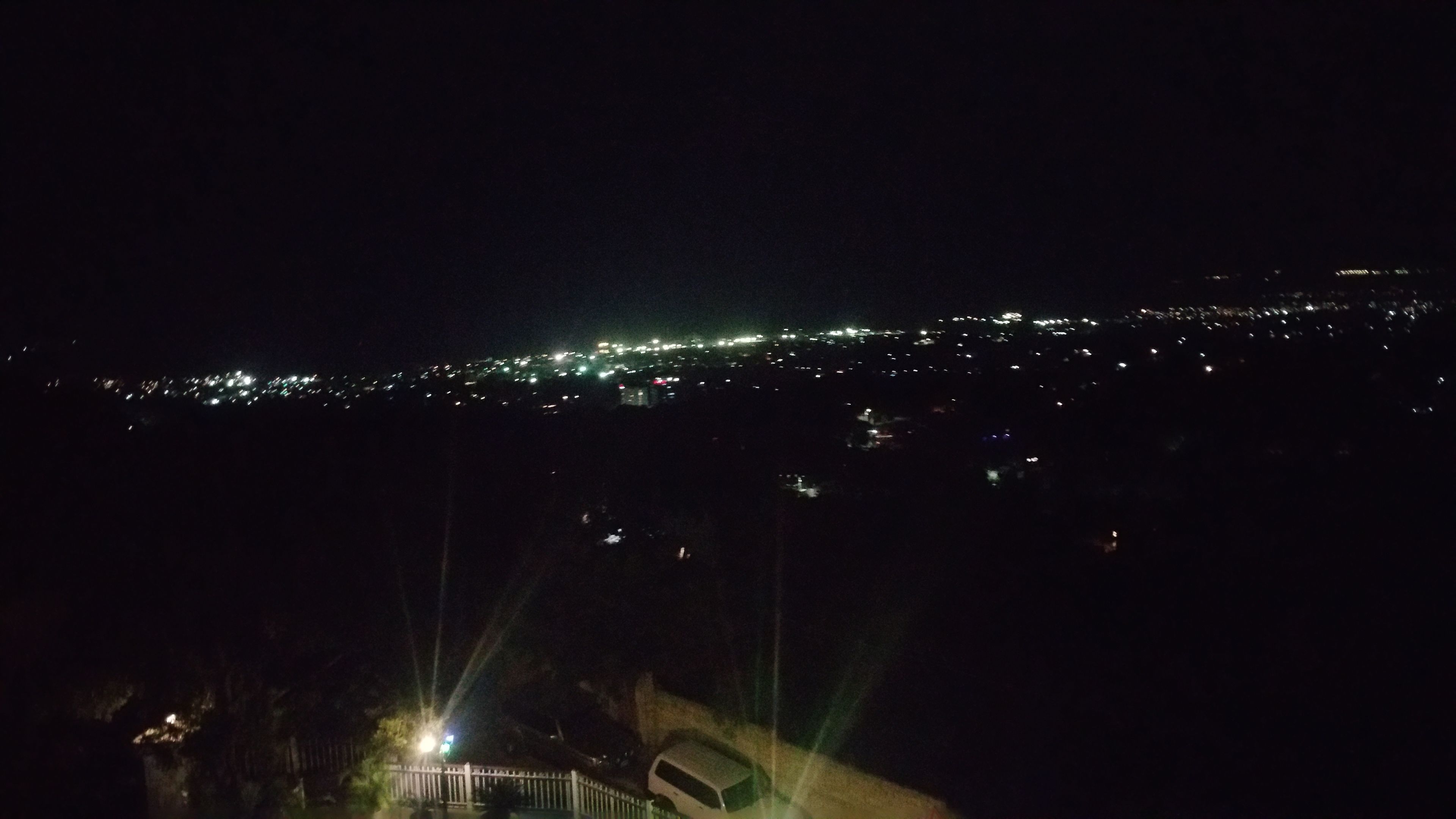 Ein Nachtblick auf eine Stadtlandschaft mit ferner Stadtansicht, einem Zaun und einem Fahrzeug.