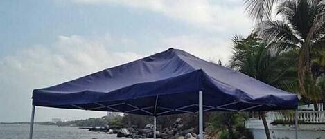 A beachside setup with a blue canopy, white plastic chairs, and a table.
