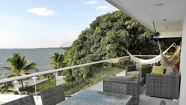 Een balkon met een tafel en stoelen met een glazen blad, met uitzicht op een waterpartij en een weelderig groen gebied.
