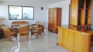 Een kamer met een eettafel, stoelen, een houten kast en een groot raam met uitzicht op groen.