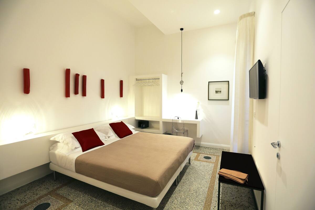 A modern hotel room with a bed, bedside tables, a TV, and a bathroom visible in the background.