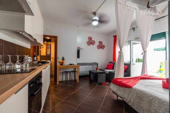 A compact hotel room with a kitchenette, a bed with a canopy, a dining area, and a small TV.