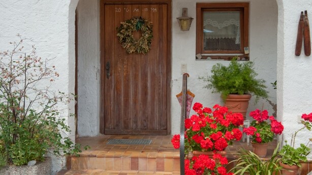 Un edificio de paredes blancas con una puerta de madera, una ventana y una maceta con flores rojas.