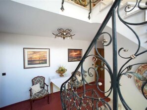 Una escalera con barandillas metálicas decorativas que conduce a un piso superior. En la pared hay dos cuadros enmarcados, una silla y una mesa con un jarrón de flores.