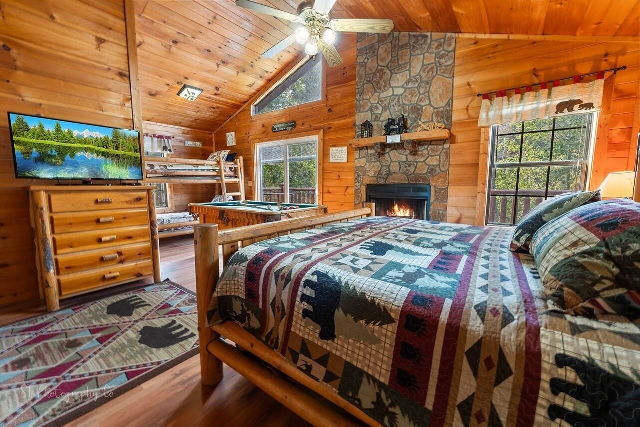 A cozy cabin interior with a bed, wooden dresser, television, fireplace, and a pool table.