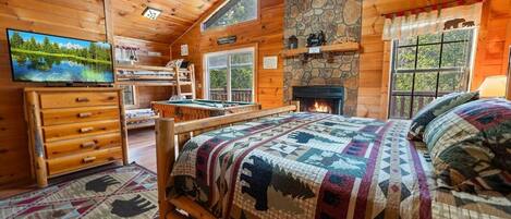A cozy cabin interior with a bed, wooden dresser, television, fireplace, and a pool table.