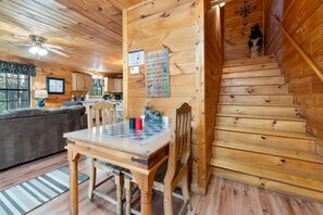 A cabin interior with a wooden staircase, a dining table with chairs, a sofa, and a kitchen area.