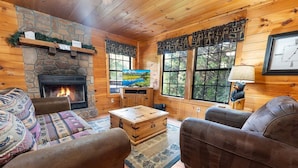A cozy cabin interior with a stone fireplace, wooden furniture, and large windows offering a view of the outdoors. - Paw Prints (Sevierville)