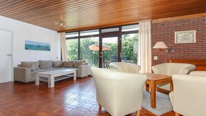 A spacious living room with a wooden ceiling, large windows, and a view of greenery outside. - Holiday house in Taddigshörn 303 - holiday houses beach castles (Esens)