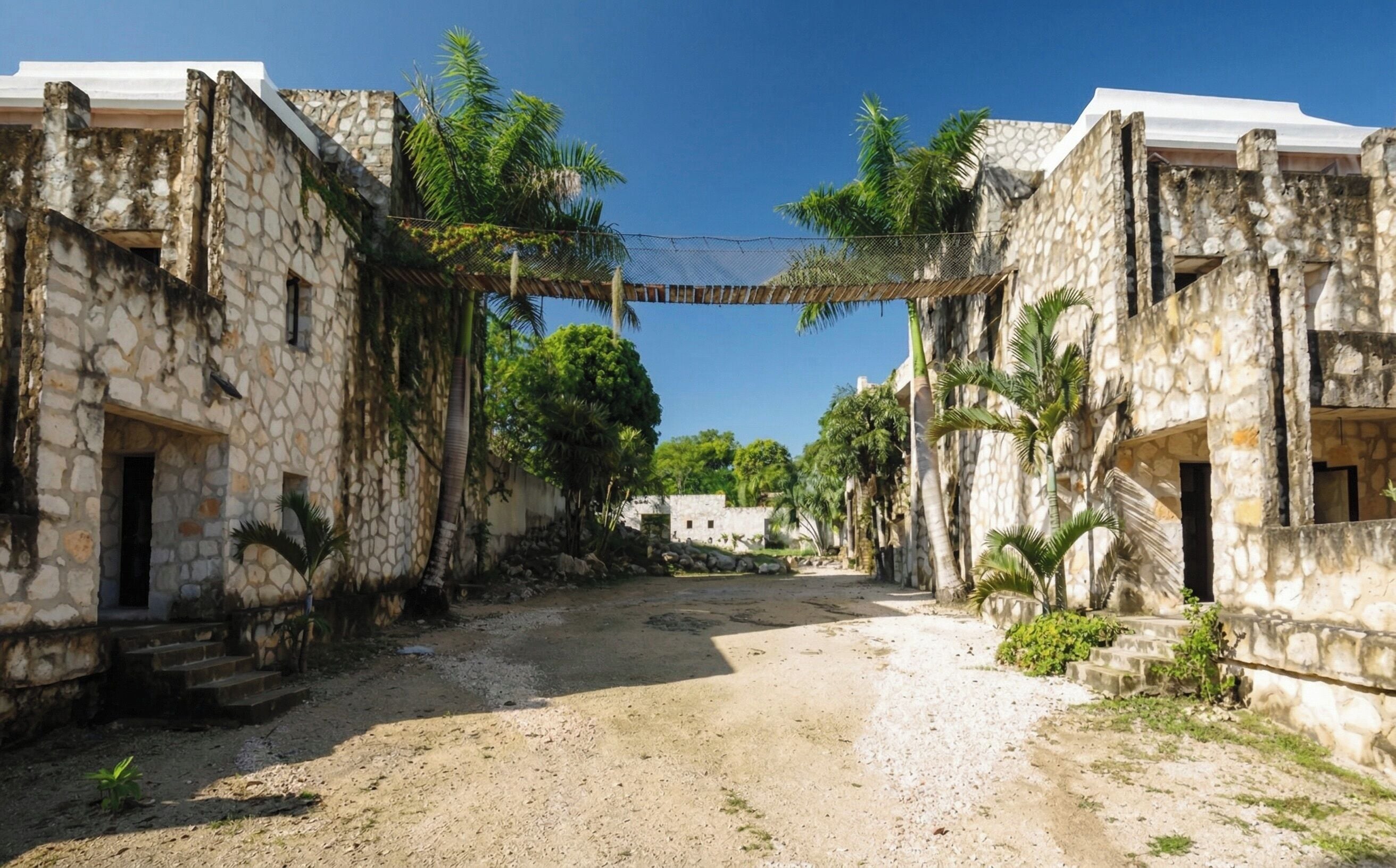A stone building with a dirt path, palm trees, and a clear sky.