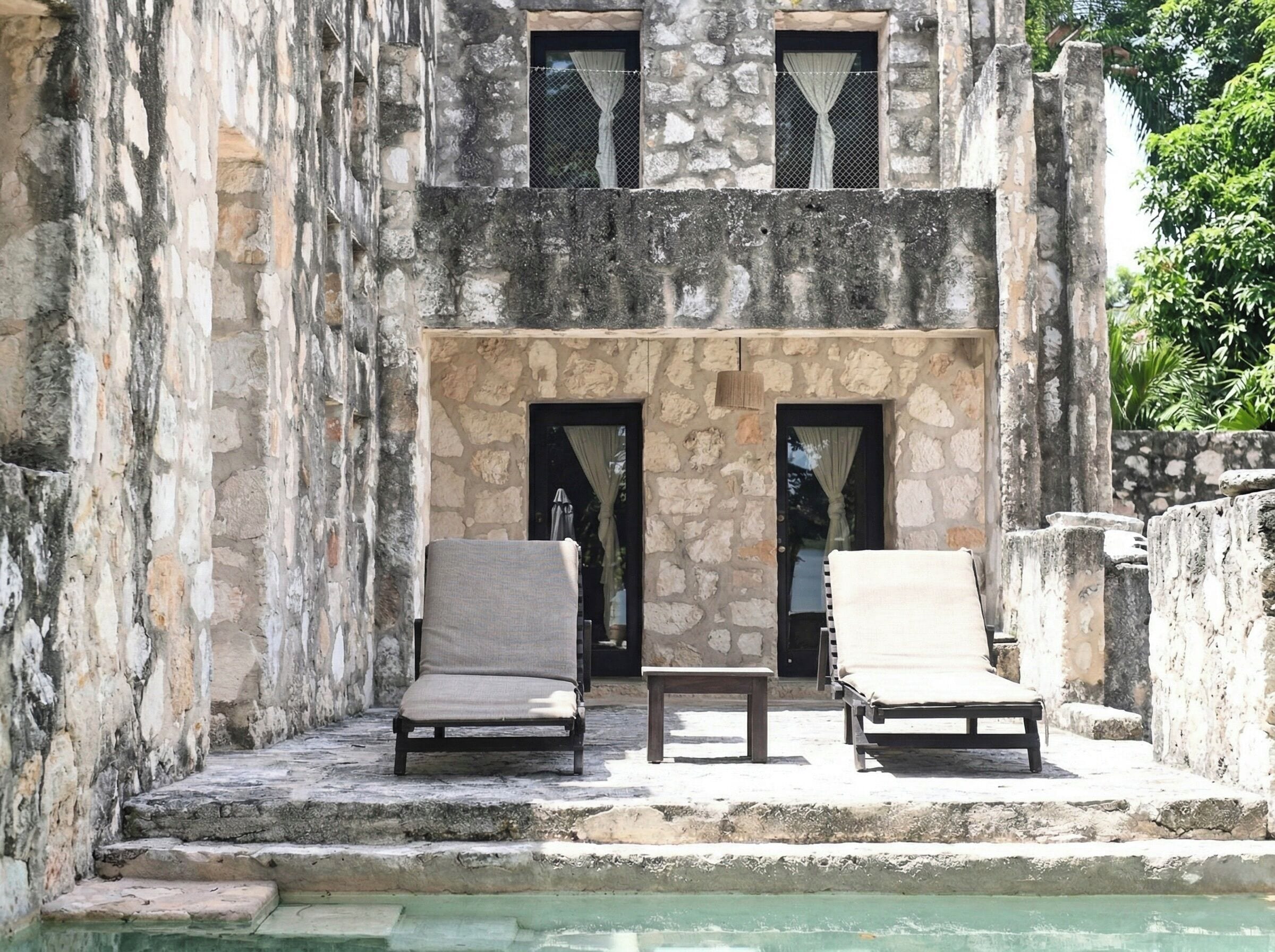 Un edificio de piedra con dos puertas, una piscina y dos sillones de descanso.
