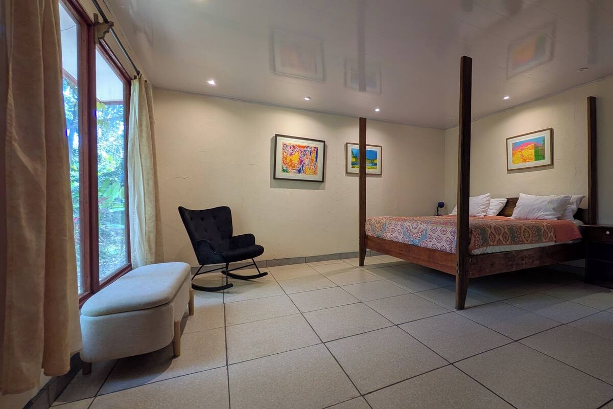 Een slaapkamer met een hemelbed, een schommelstoel, een bankje en twee ingelijste kunstwerken aan de muur.
