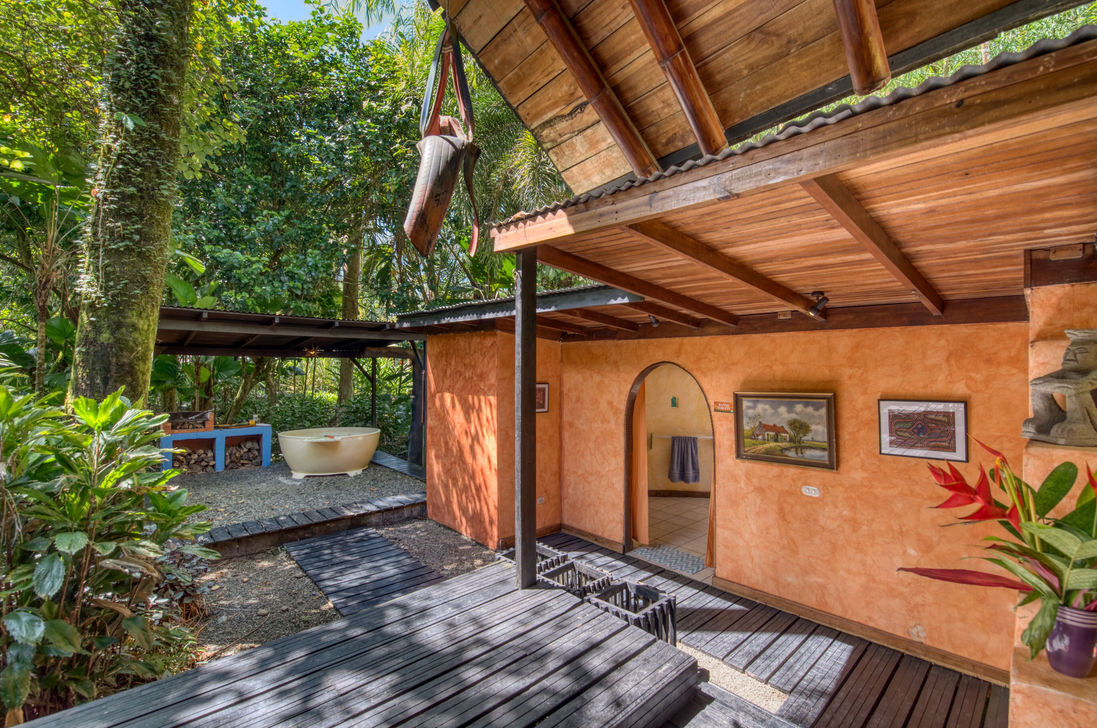 A wooden deck with a covered area, surrounded by lush greenery and a rustic building with a tiled floor and a hanging pot.