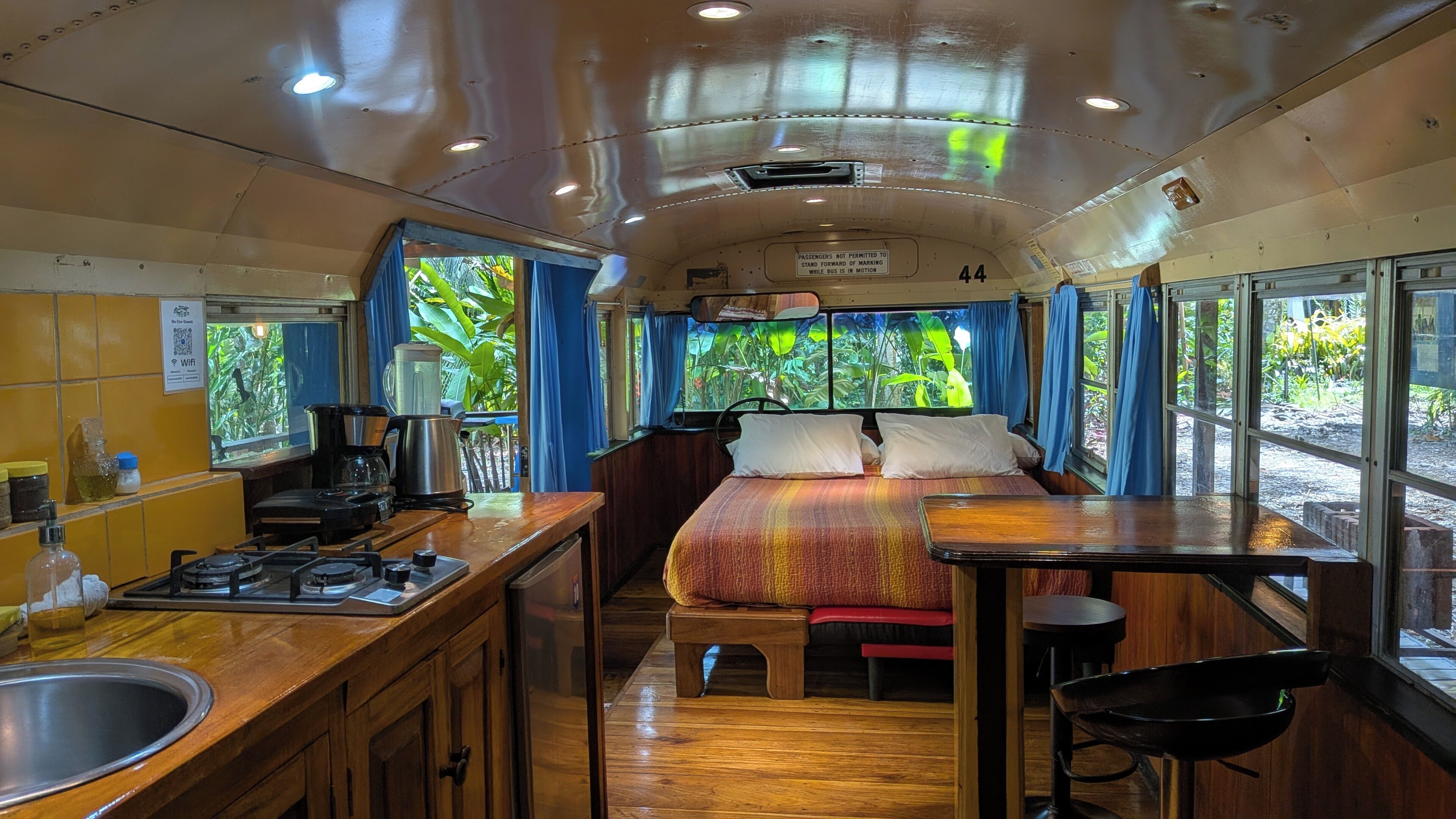 A compact, well-lit train car with a kitchenette, a bed, and large windows offering a view of greenery.