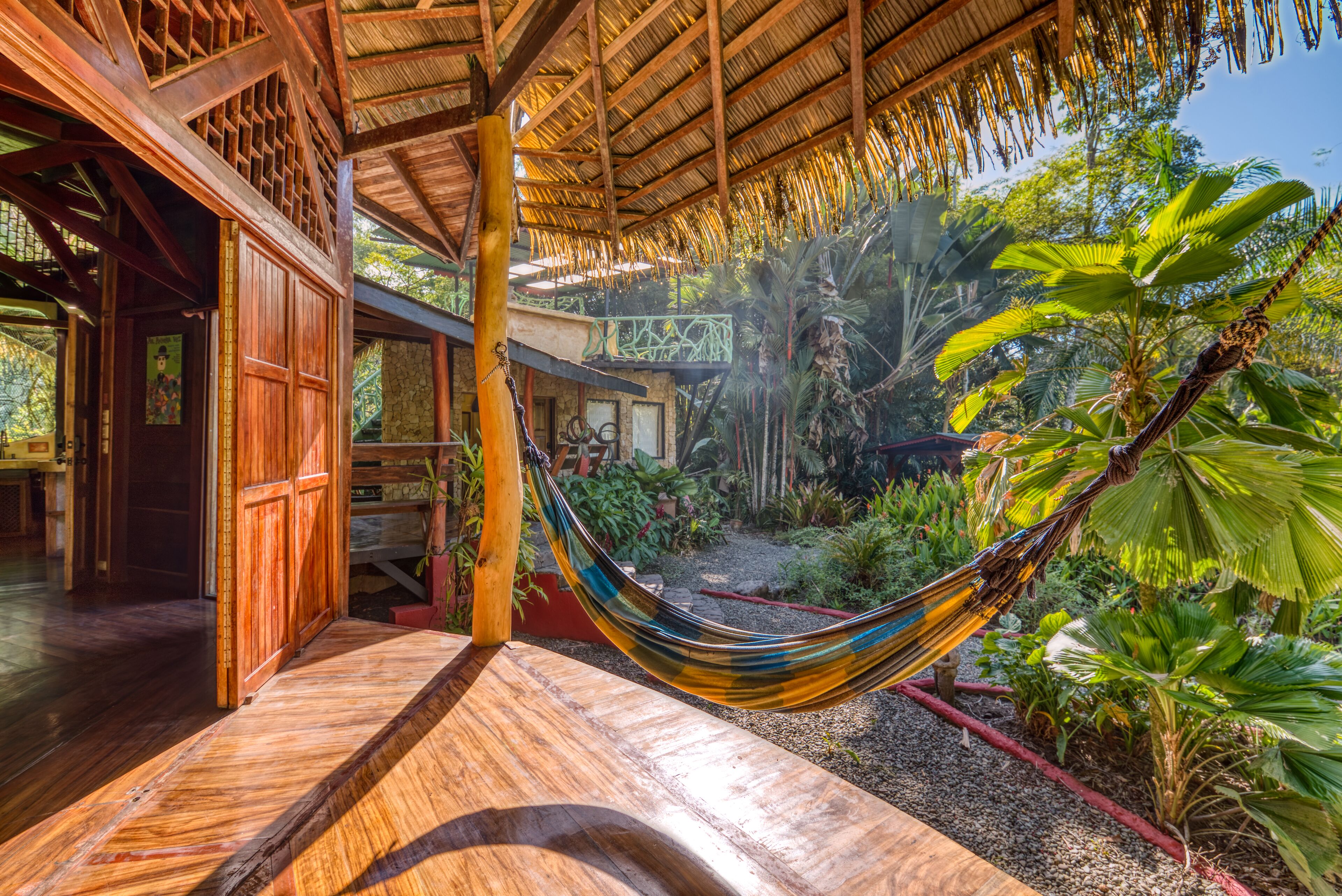 A hammock in a wooden outdoor area with a thatched roof, surrounded by lush greenery and a stone pathway.