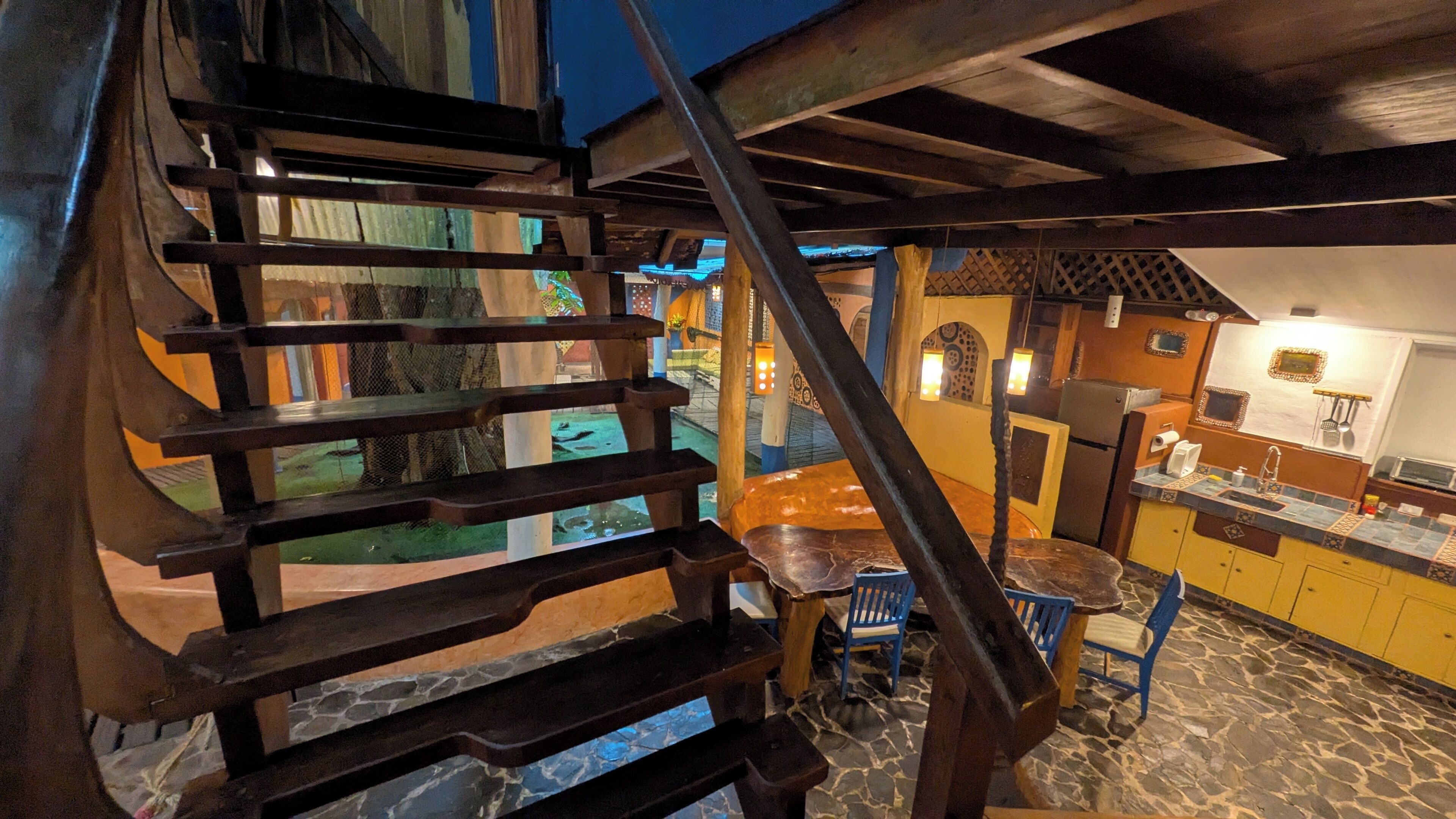 Un escalier en colimaçon mène à une salle à manger dont le sol est en pierre, et on aperçoit une cuisine au fond.
