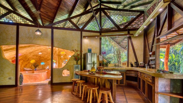 Eine geräumige Küche mit einer großen Kochinsel, Holzstühlen und Blick auf einen üppigen Garten durch eine Glaswand.