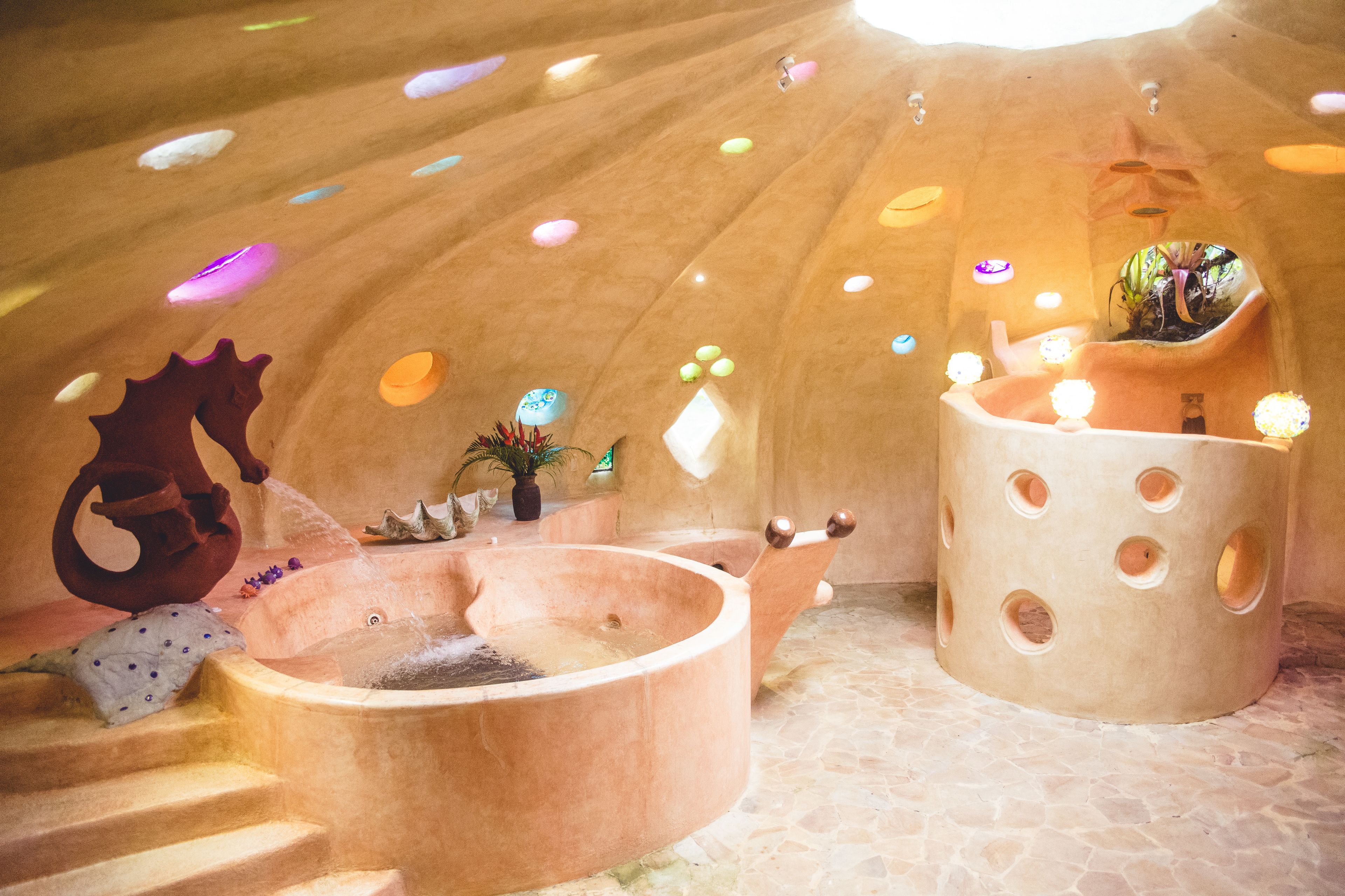 Una habitación circular de piedra con una fuente central, un objeto cilíndrico grande con agujeros y una escalera que conduce a un nivel superior.