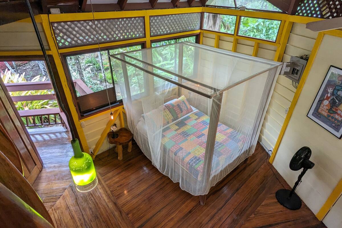 Een kamer met een bed onder een klamboe, houten vloeren, een klein krukje, een tafellamp en een raam met uitzicht op groen.