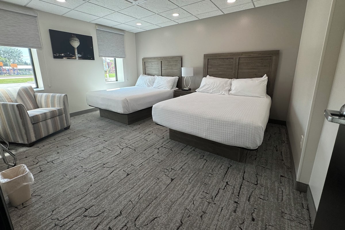 A hotel room with two beds, a chair, a trash bin, and a window with blinds.