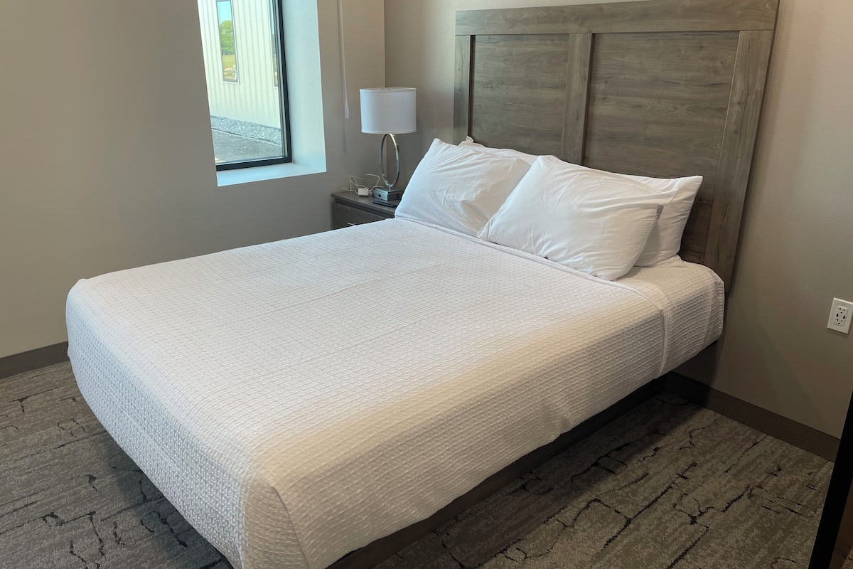 A neatly made bed with white linens in a room with a wooden headboard, a bedside lamp, and a window with a view of a building outside.